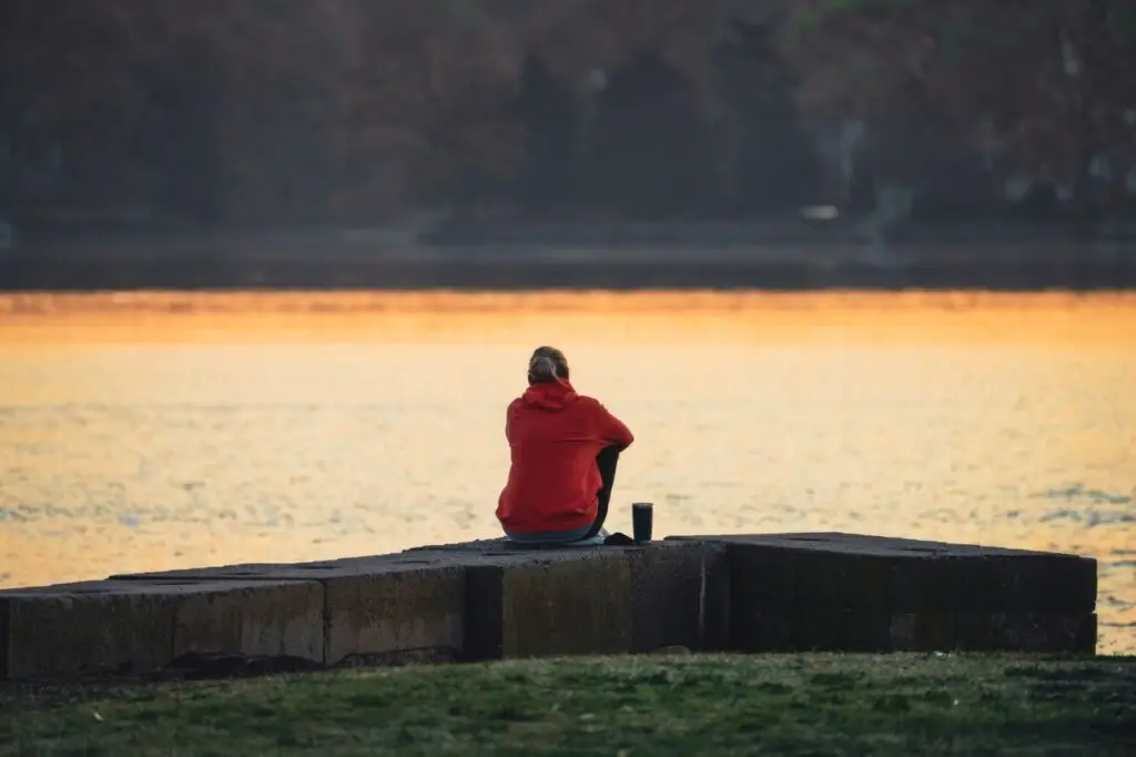 Person in red hoodie sitting by a lake at sunset, reflecting in serene solitude.
